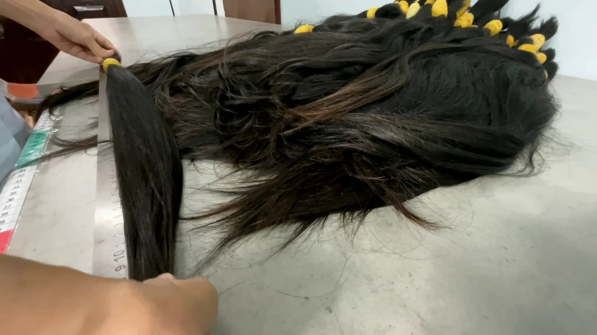 A hand lifting one dark ponytail bundle from a pile of donated hair on the intake table